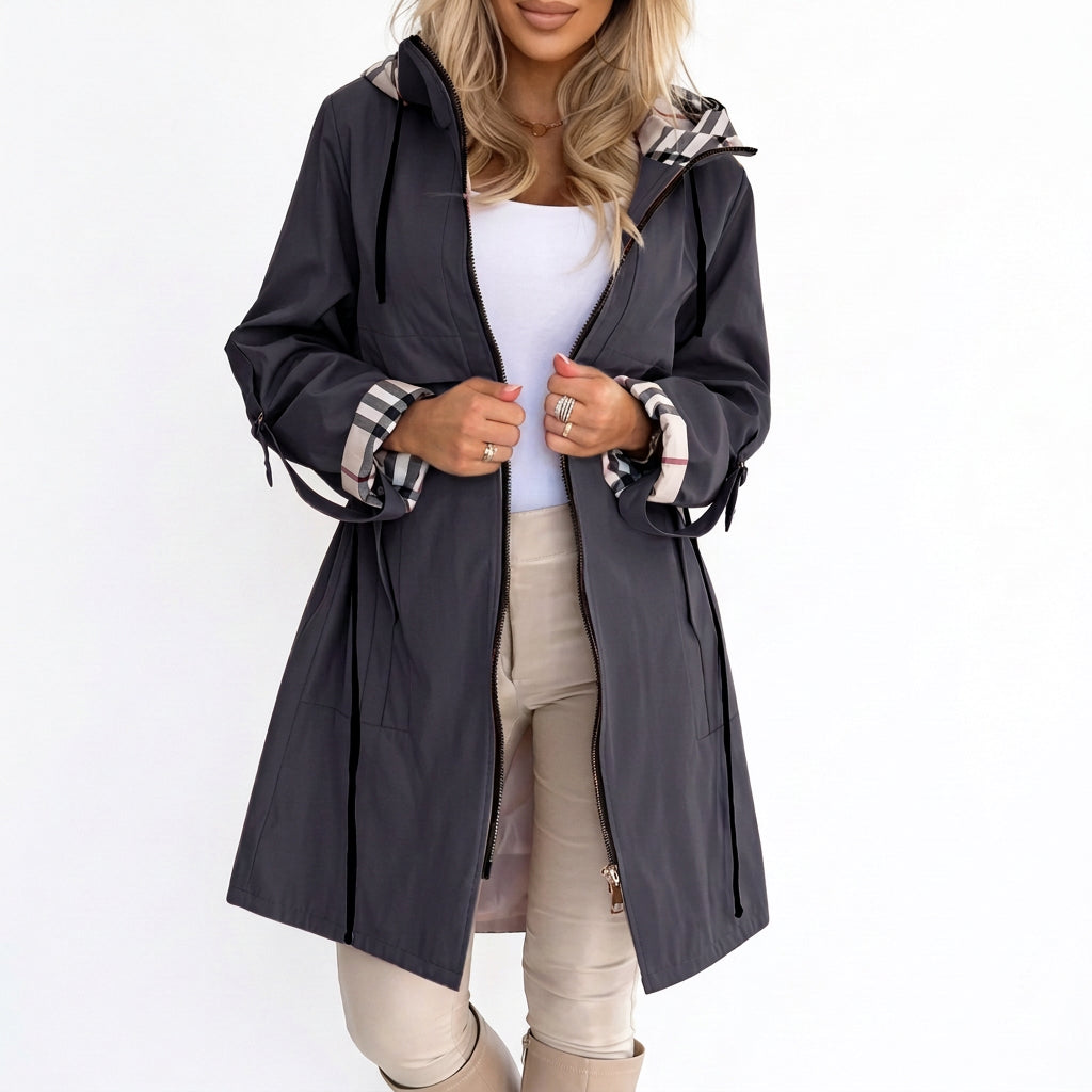 Dark grey hooded coat with plaid lining on cuffs and hood, worn over white top and beige pants.