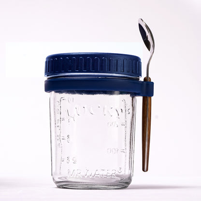Glass overnight oats jar with blue lid, measurement markings, and a spoon held in a side silicone loop