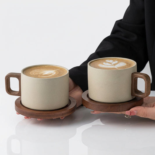 A person holding two minimalist stoneware mugs with wooden handles and matching wooden coasters filled with latte art coffee.