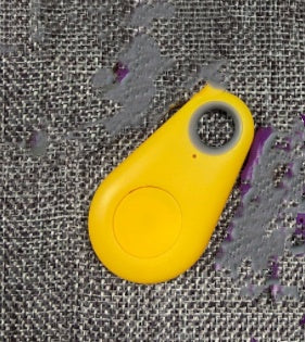 Yellow teardrop-shaped waterproof Bluetooth tracker with a center button on a textured grey fabric background.