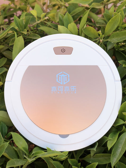White robotic vacuum with rose gold lid and power button resting on green foliage.