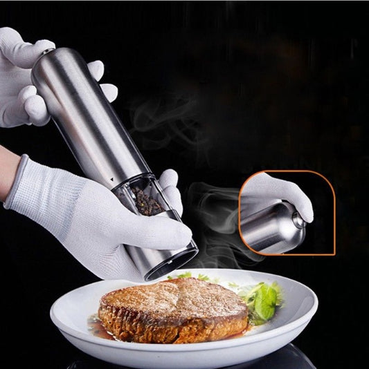 gloved hands operating a stainless steel electric pepper grinder over a plate of cooked steak