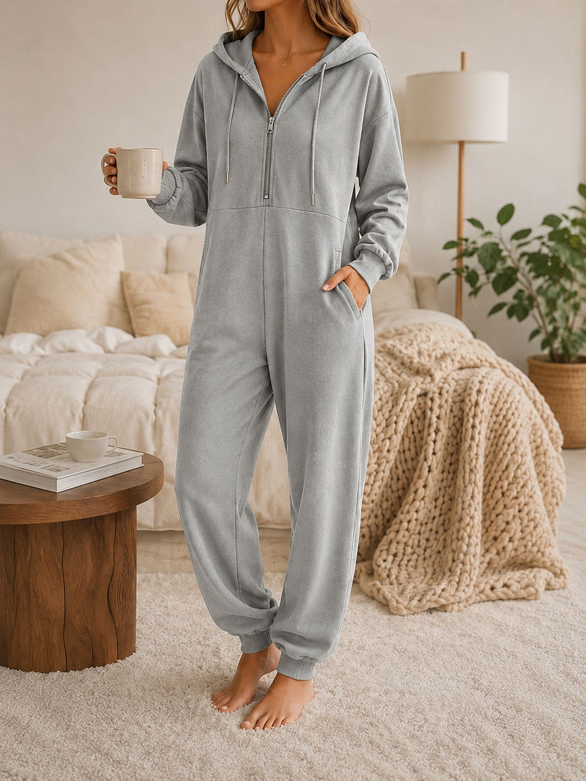 Model in a hooded zip-up jumpsuit holding a ceramic mug, standing by a sofa and side table