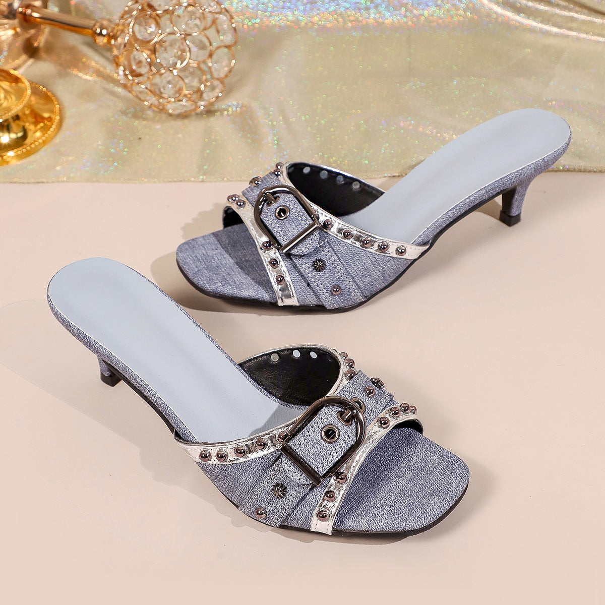 Pair of blue denim slide sandals with kitten heels, decorative buckles, and metallic rivet trim.