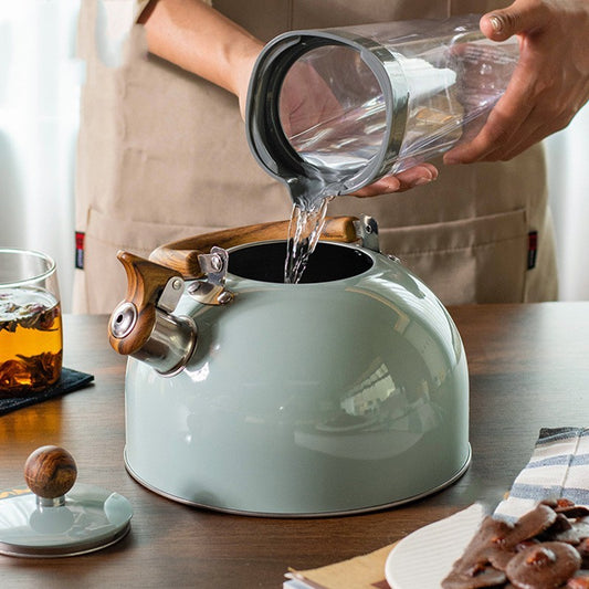 Water pouring from a clear pitcher into a sage green tea kettle with a wooden handle on a wooden table.