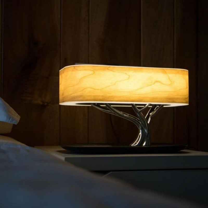 Modern table lamp with a glowing rectangular wood-grain shade supported by a silver tree-branch base on a nightstand.
