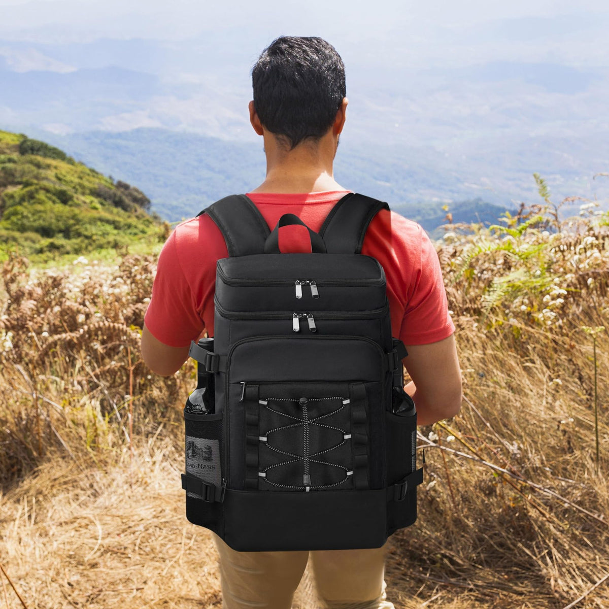 Man wearing a black cooler backpack standing outdoors facing a mountainous landscape.