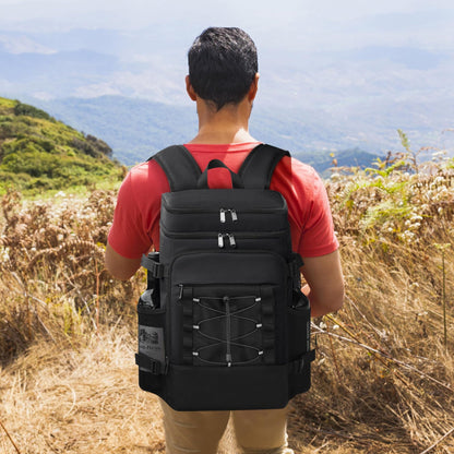Man wearing a black cooler backpack standing outdoors facing a mountainous landscape.