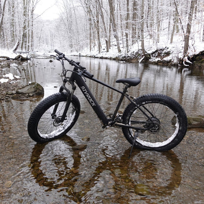Black aluminum frame all-terrain bike with fat tires in a snowy stream for winter trail riding.