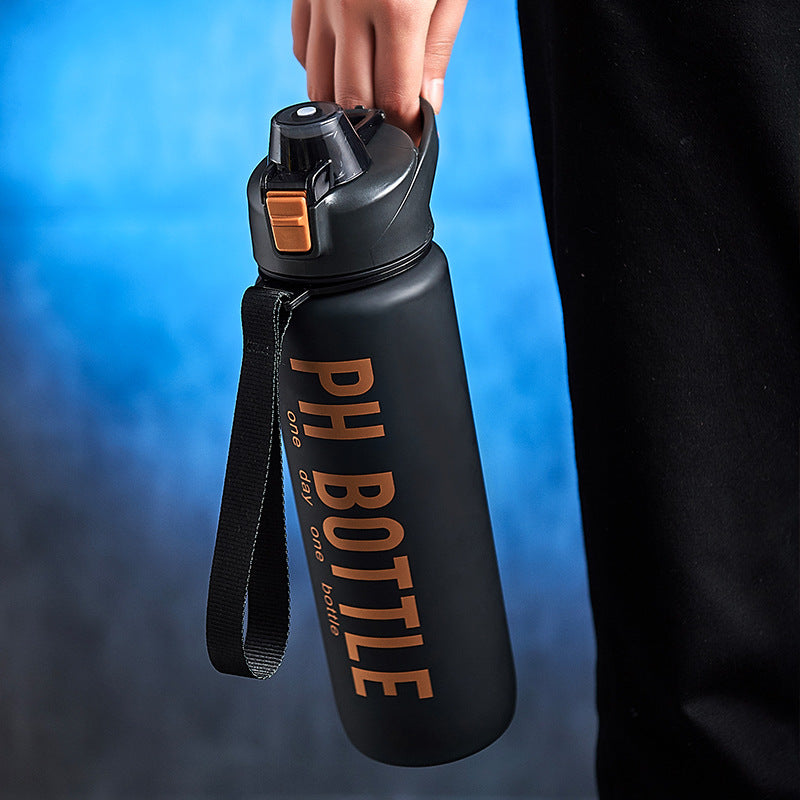 Person holding a matte black water bottle with a black carrying strap, orange lid latch, and large orange text.