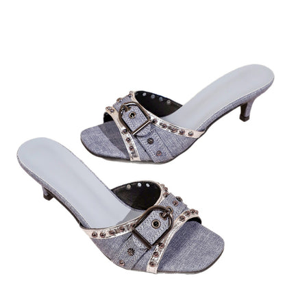 Pair of blue denim slide sandals featuring silver-toned buckles, studded trim, and kitten heels against a white background.