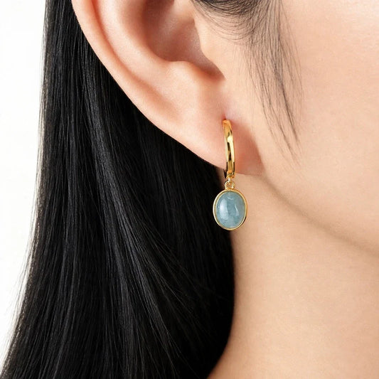 Gold hoop earring with a dangling light blue oval stone worn on an earlobe.