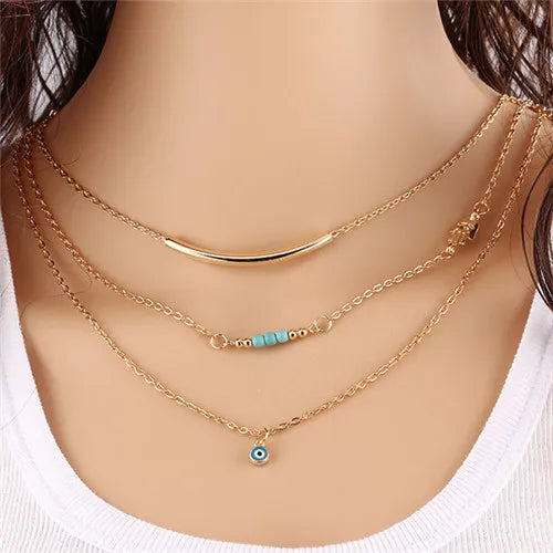 Gold-colored multilayer chain necklace with a curved bar, turquoise beads, and an evil eye pendant.
