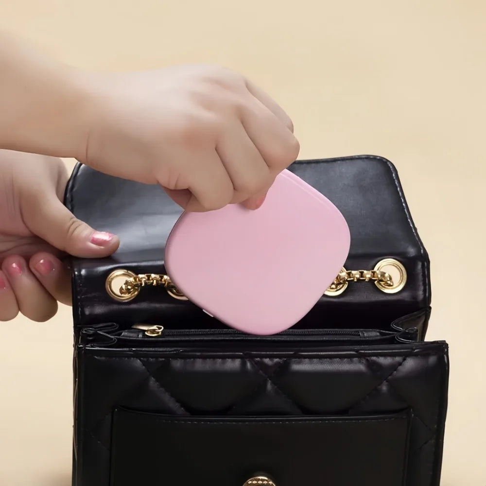 Round light pink LED makeup mirror with adjustable brightness for travel, being placed into a black quilted handbag.