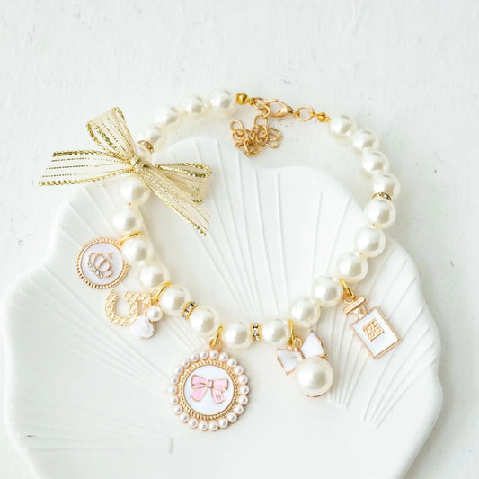 White pearl pet collar with gold bow, charms, and rhinestone accents on a white shell-shaped dish.
