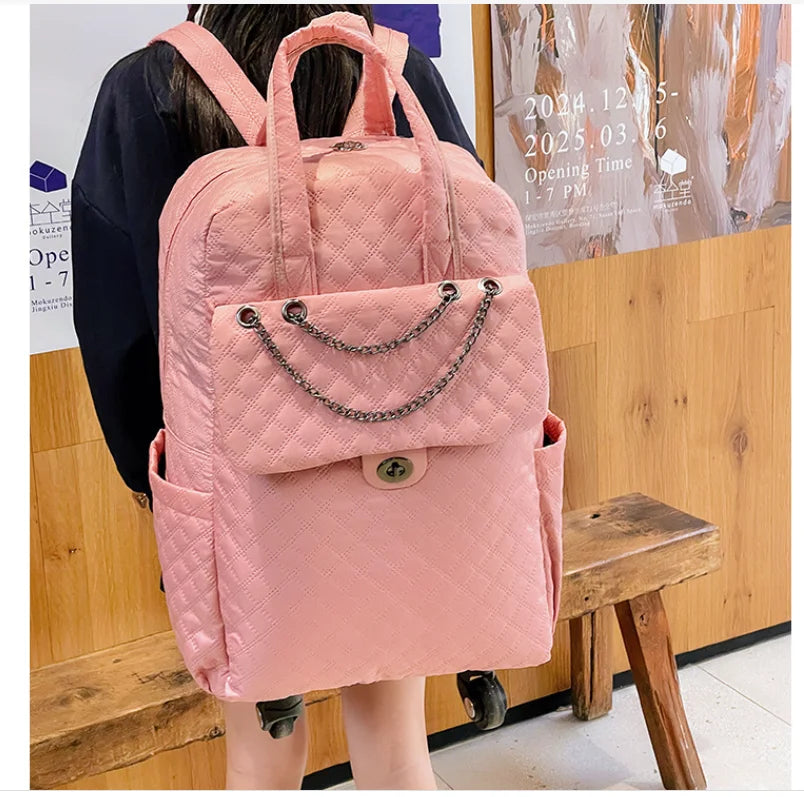 Pink quilted nylon rolling backpack with top handles, front flap pocket, chain detail, and wheels.