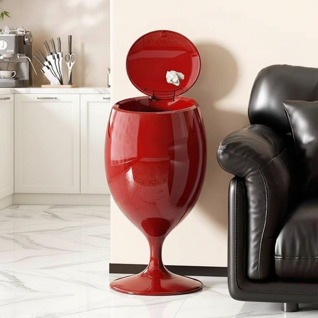 Red wine-glass-shaped trash can with open lid, standing on wooden floor next to a black leather sofa.