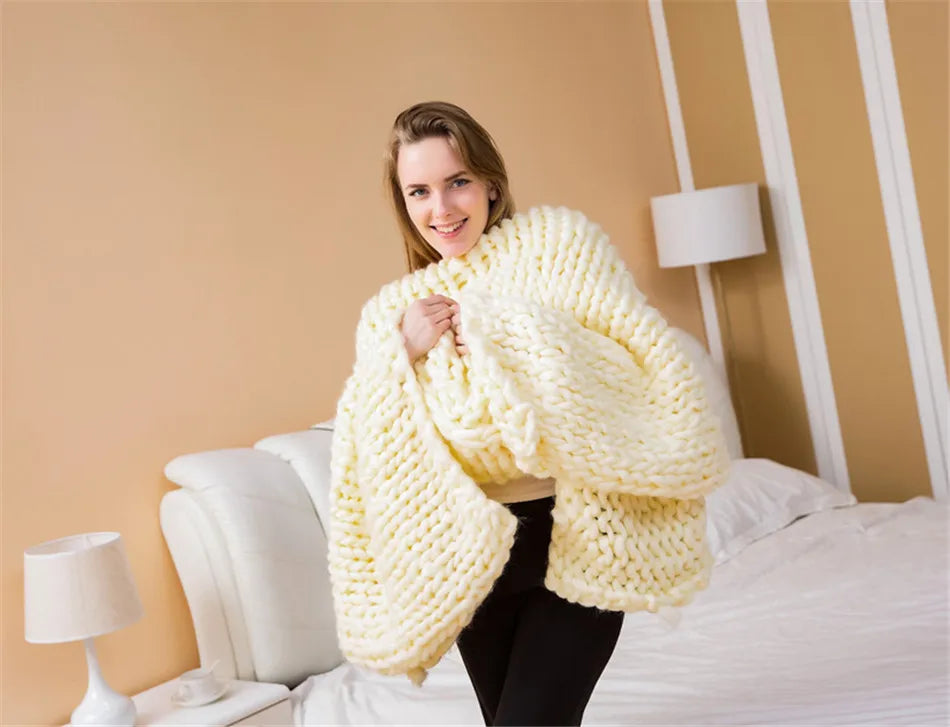 A woman wraps herself in a chunky, cream-colored, hand-knitted yarn blanket in a bedroom.