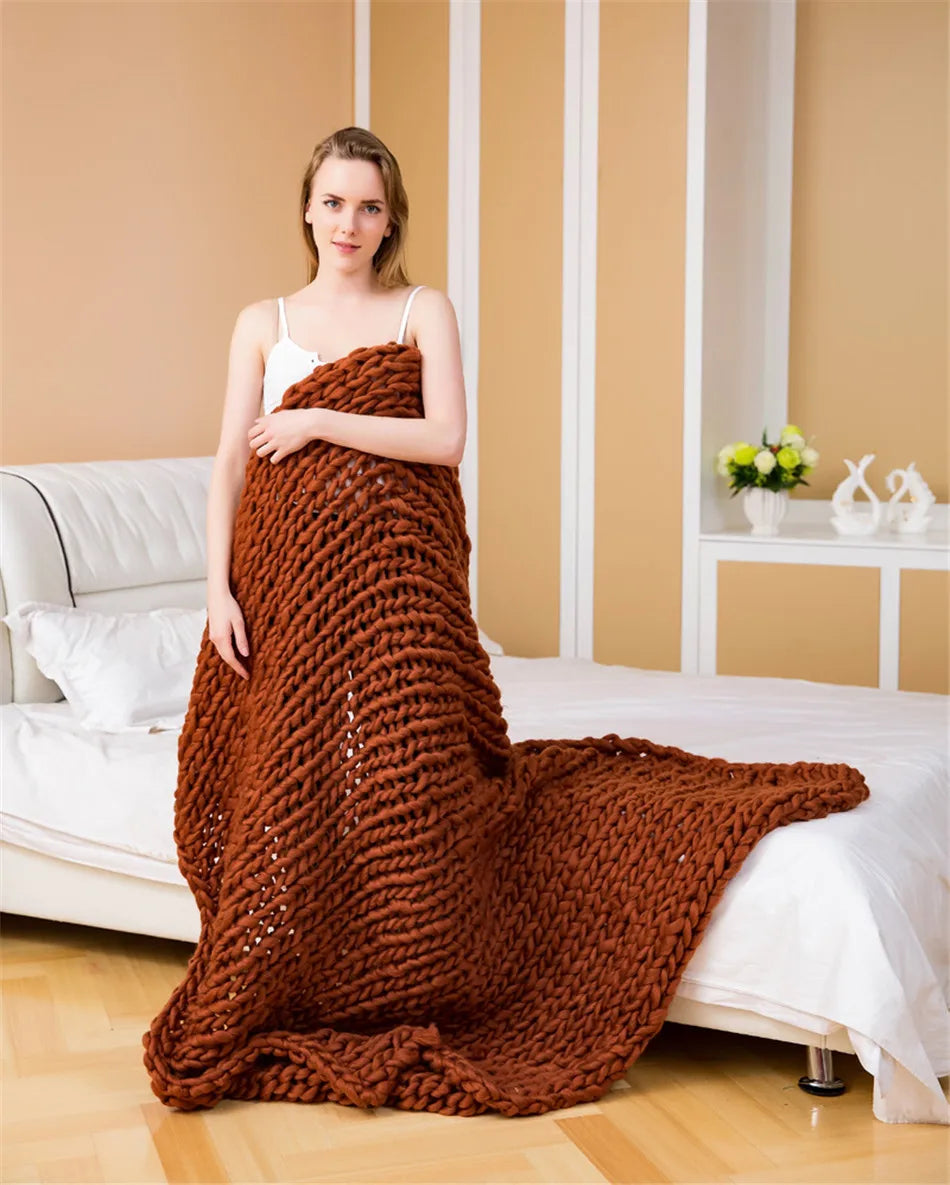 A woman wrapped in a chunky, rust-orange hand-knitted yarn blanket, standing in a bedroom.