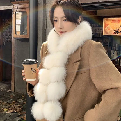 White faux fur scarf with soft pom-pom segments, worn wrapped around the neck by a woman in a brown coat.