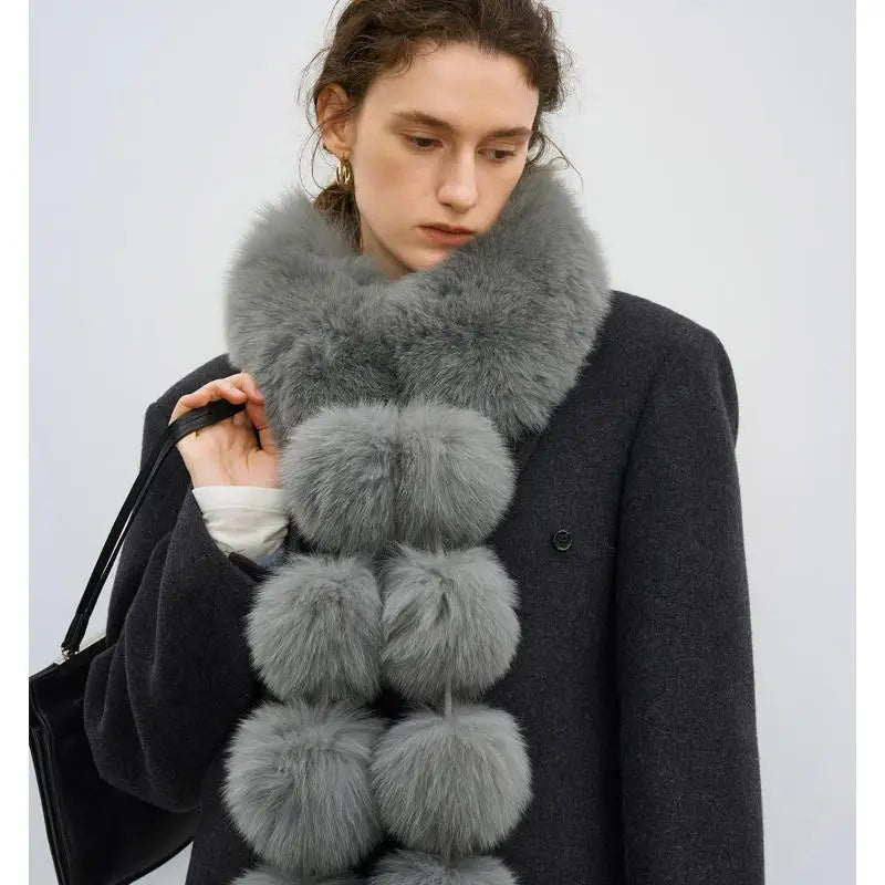 A woman wears a long, gray faux fur scarf with large pom-pom details and a dark coat.