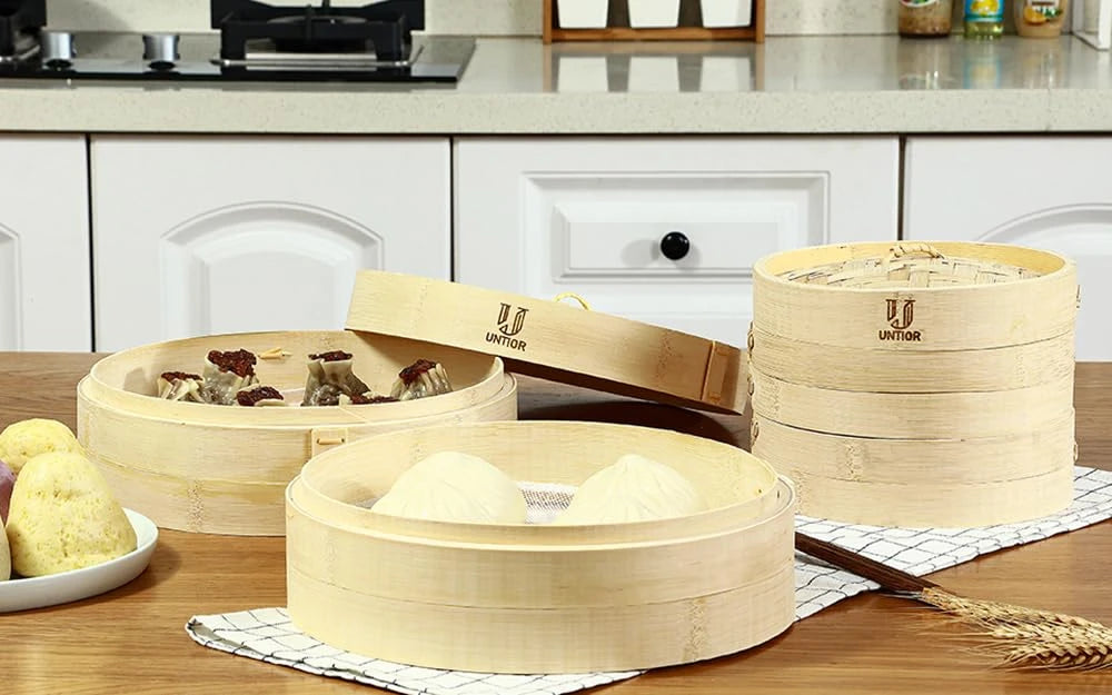 Round bamboo dumpling steamers with lids, holding bao buns and dumplings, on a kitchen table.