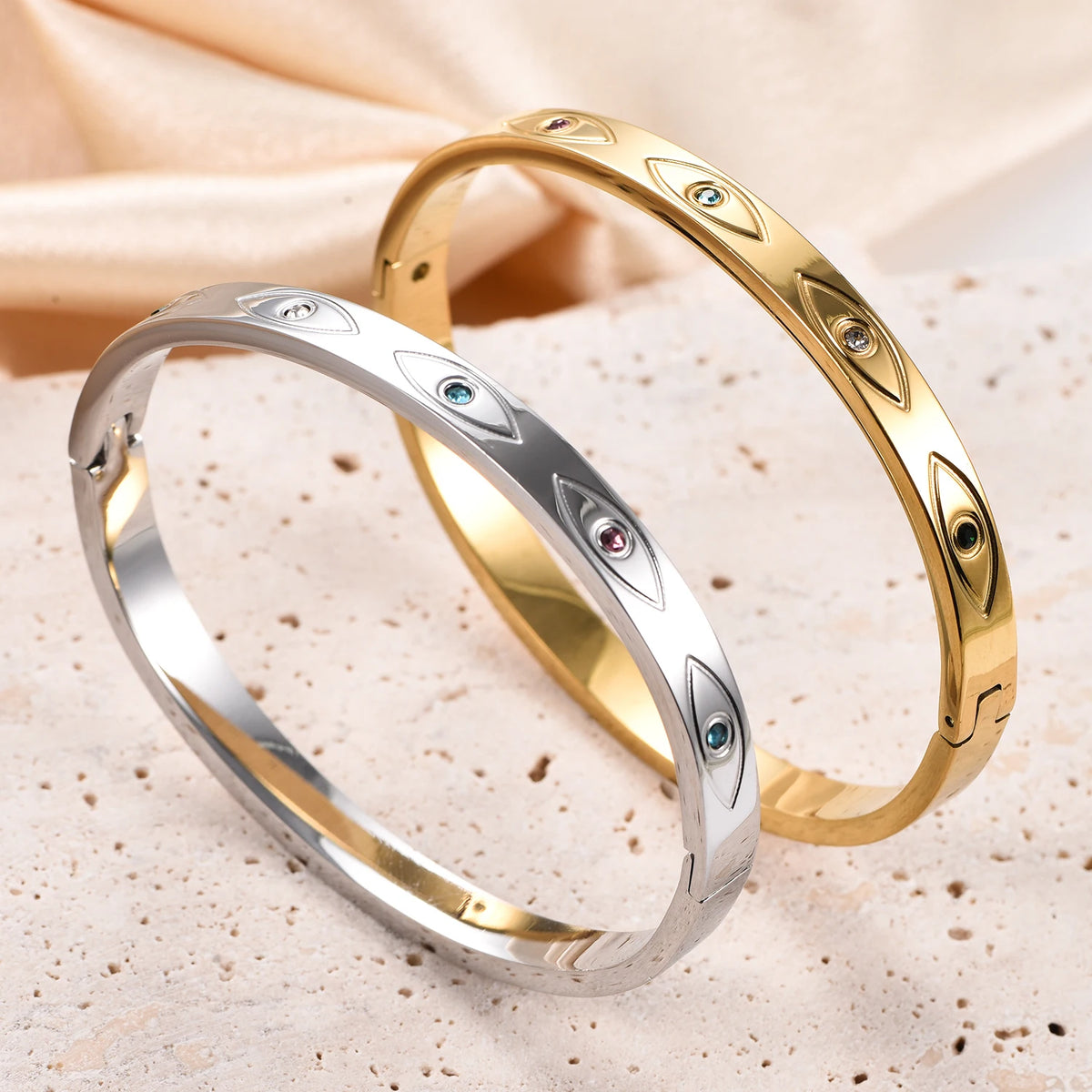 Two stainless steel cuff bangles, one silver and one gold, engraved with evil eye motifs set with colorful crystals.