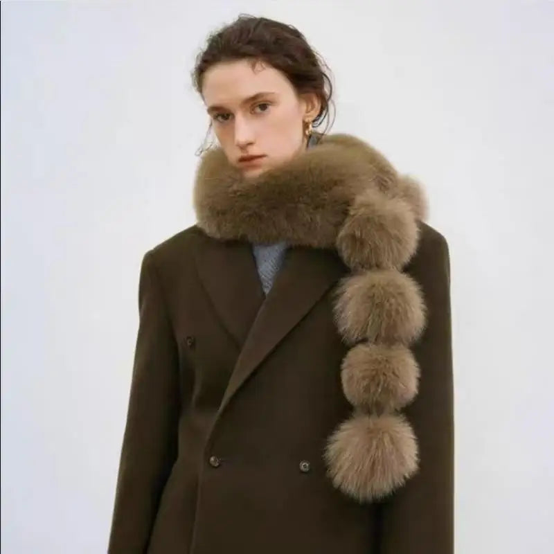 Brown faux fox fur scarf with oversized pom-pom accents, worn around the neck over a dark blazer.