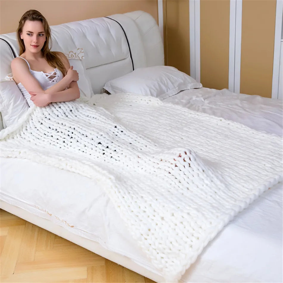 A woman sits on a bed, covered in a white chunky knit yarn blanket.