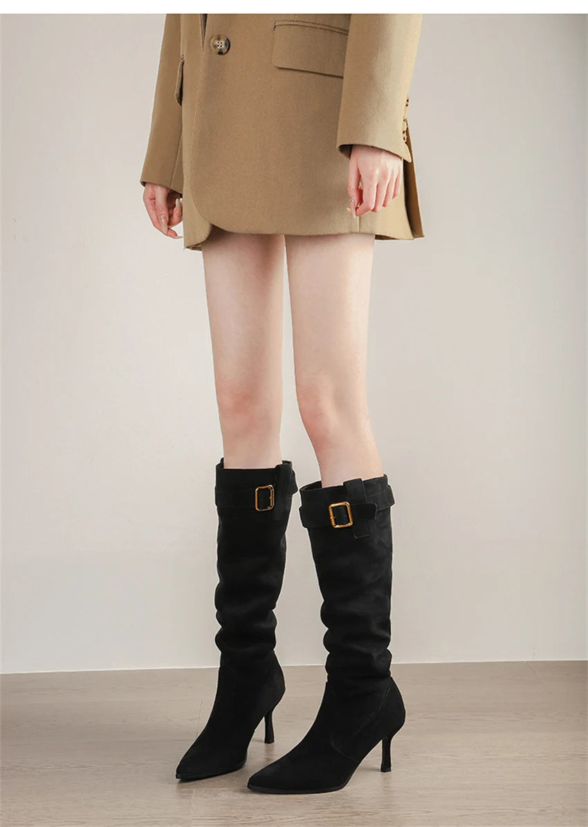 Black suede pointed-toe knee-high boots with thin high heels and a gold-buckled strap.