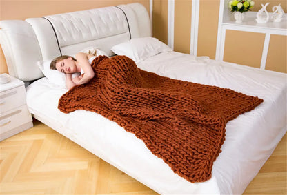 A woman sleeps under a thick, rust-brown chunky knit yarn throw blanket on a bed.