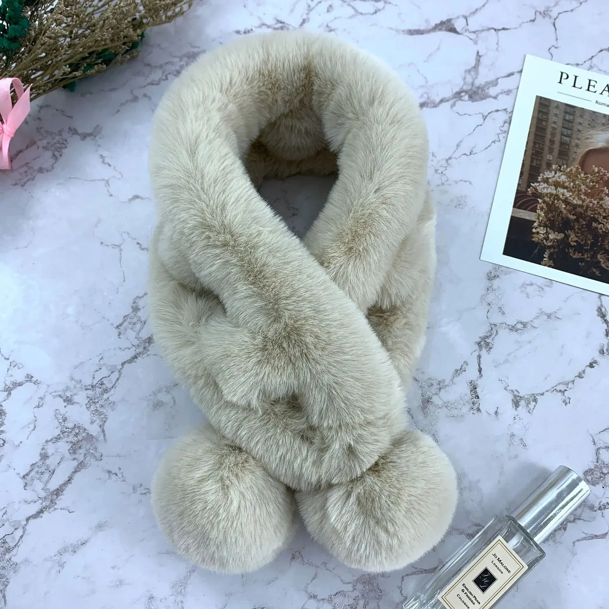 Beige faux fur crossover scarf with two fluffy pom-pom ends on a marble surface.