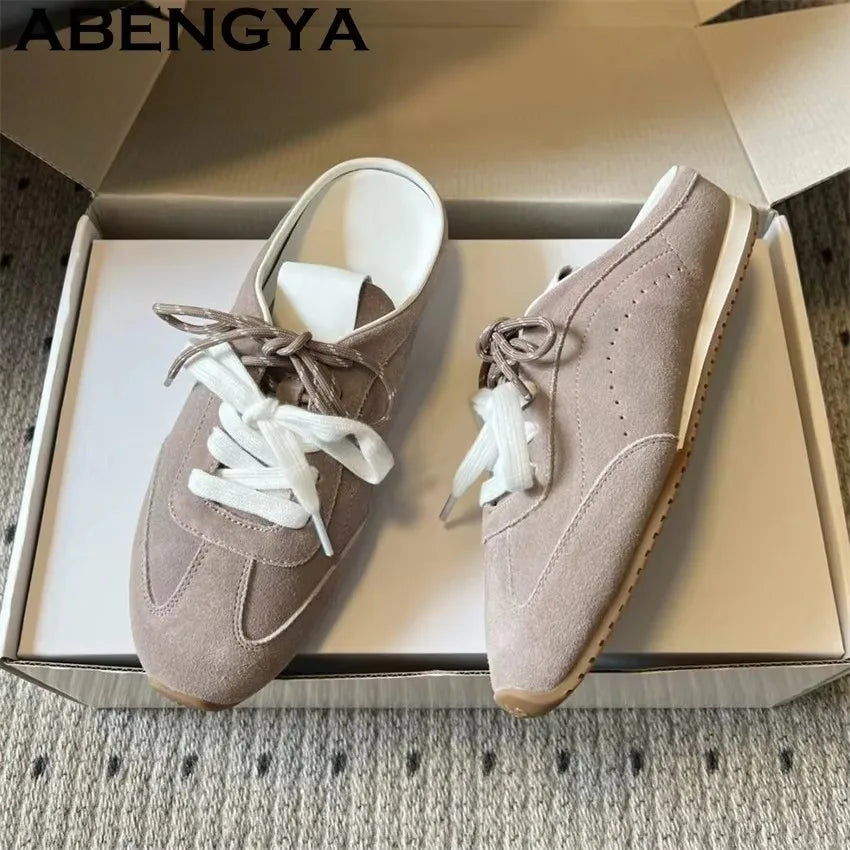 Taupe suede backless slip-on sneakers with white laces, in an open box.