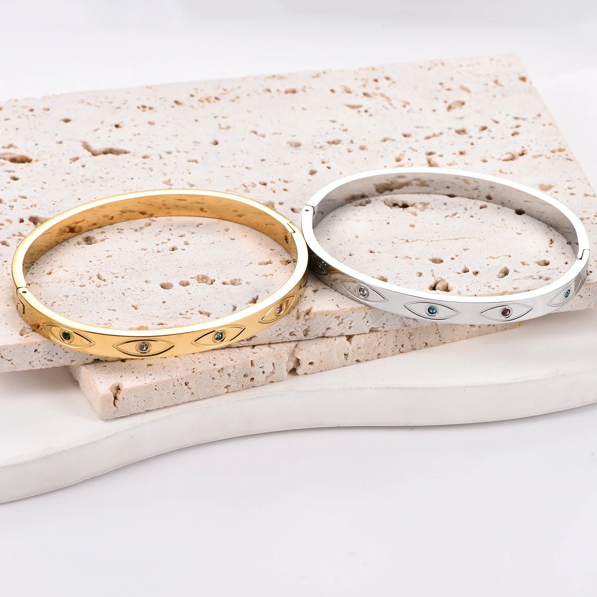 Two stainless steel cuff bangles, one gold and one silver, featuring engraved evil eye motifs with inlaid crystals.