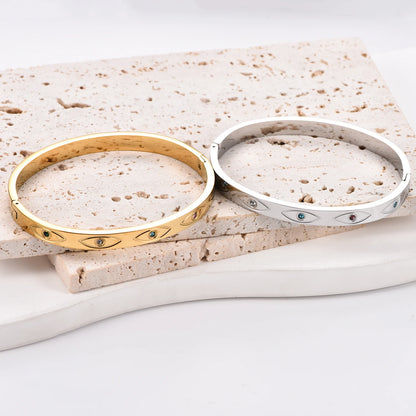 Two stainless steel cuff bangles, one gold and one silver, featuring engraved evil eye motifs with inlaid crystals.