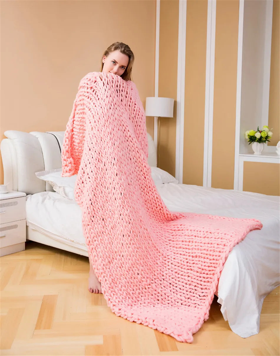 A woman wrapped in a chunky pink knit blanket, standing beside a bed with white bedding.