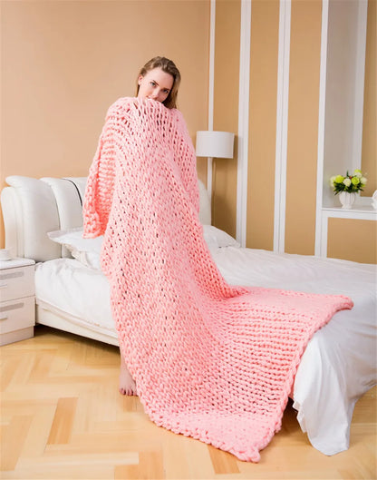 A woman wrapped in a chunky pink knit blanket, standing beside a bed with white bedding.
