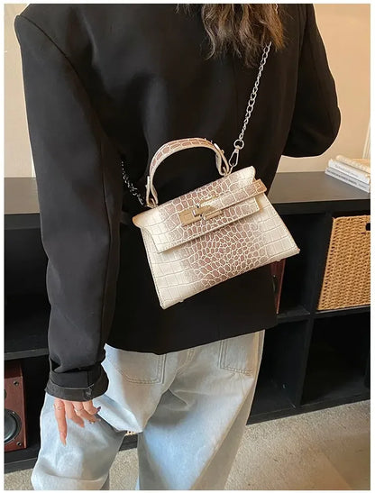 Beige faux leather crossbody diaper bag with croc pattern, handle, and chain strap for hands-free travel with baby.