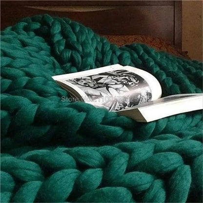 Dark green chunky hand-knitted yarn throw blanket draped on a bed with an open book.