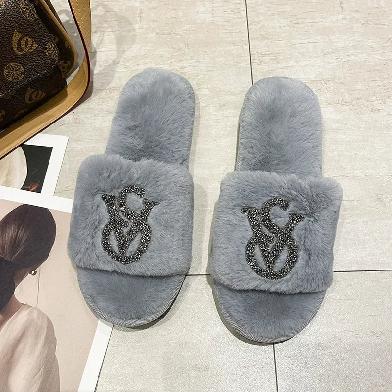 Gray faux fur slippers with rhinestone logos and non-slip soles for cozy indoor wear.