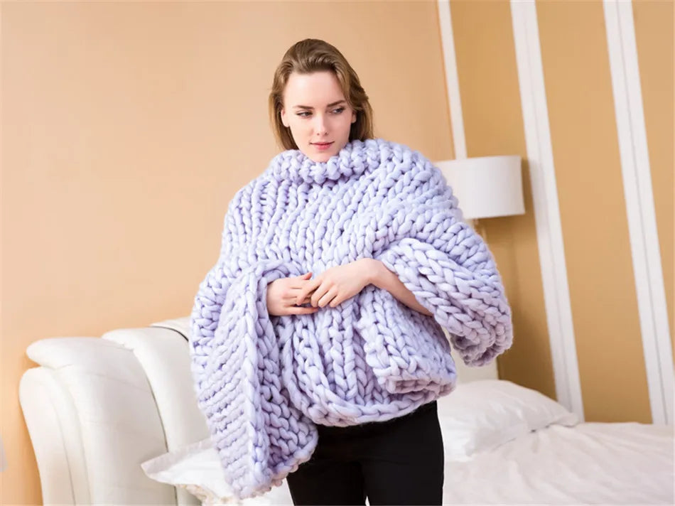 A woman wrapped in a light purple, chunky hand-knitted yarn blanket in a bedroom.