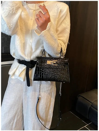 Black faux leather crossbody bag with croc texture, metal clasp, and adjustable strap for carrying baby essentials.