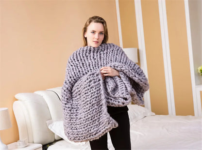 A person holds a large, chunky-knit grey yarn blanket in a bedroom.