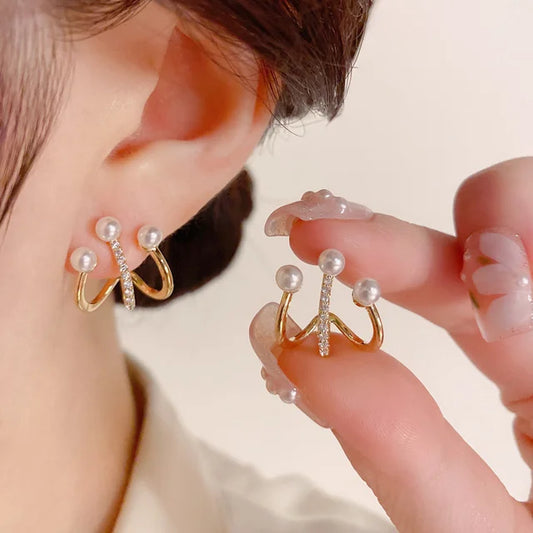 Gold stud earrings with double hoops, pearls, and a central zircon crystal, worn on an earlobe and held in fingers.