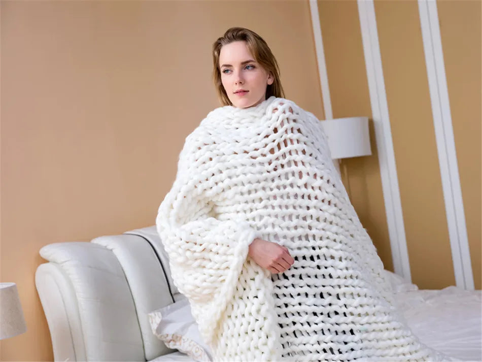 Thick white chunky hand-knitted yarn blanket draped over a person seated on a bed.