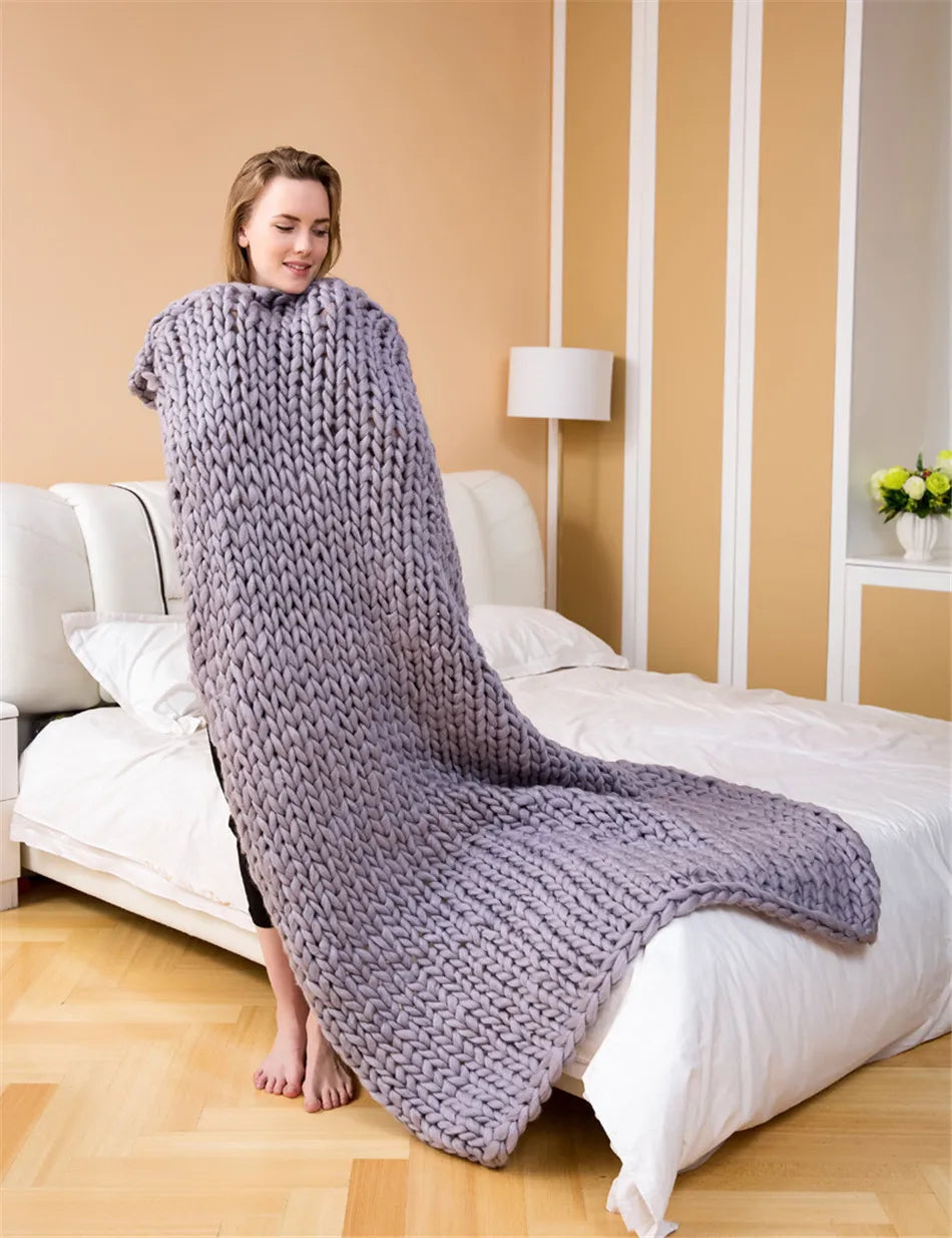 Grey chunky hand-knitted throw blanket draped in front of a person standing beside a bed.
