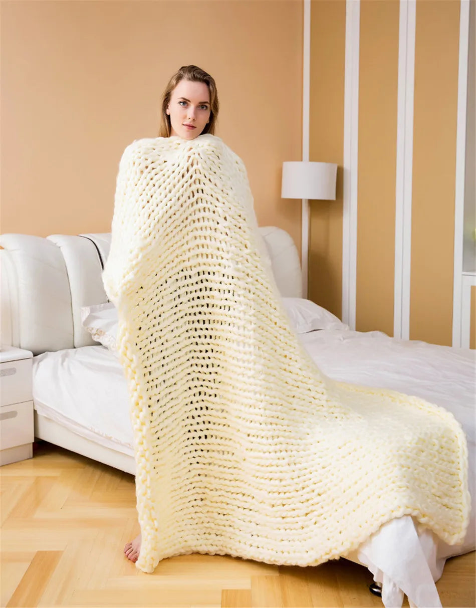 A woman wrapped in a chunky cream-colored yarn knit blanket in a bedroom.