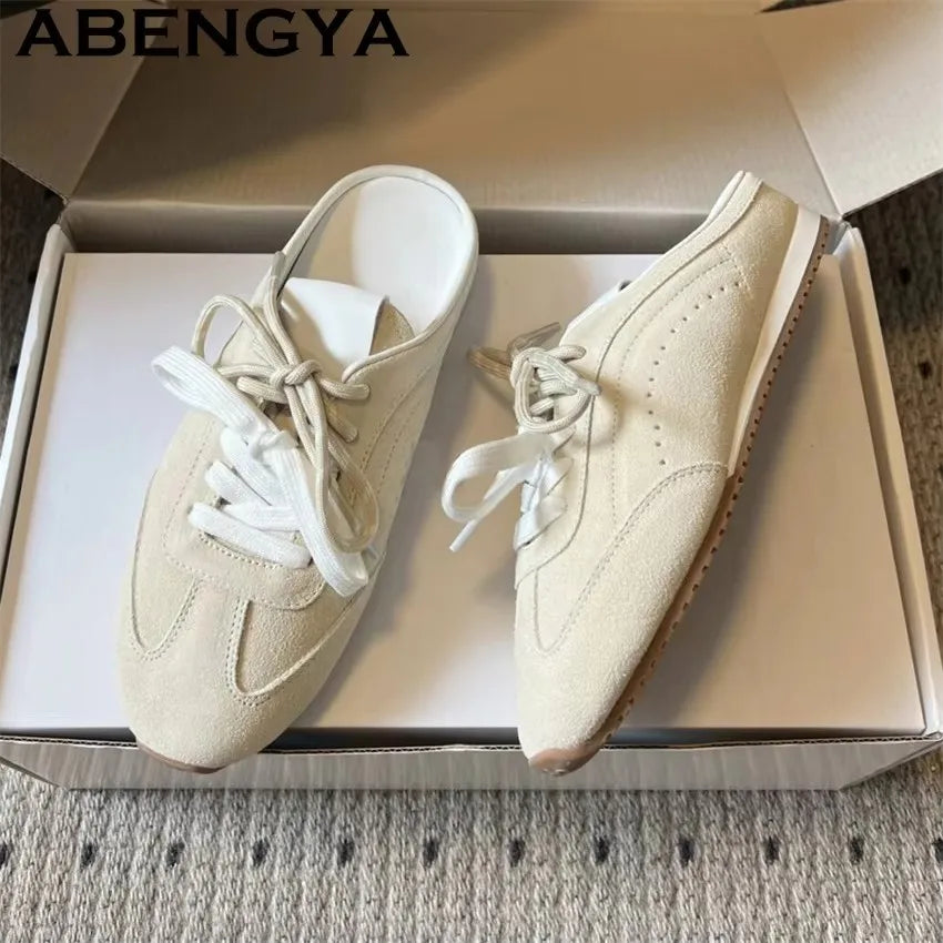 Beige suede backless slip-on sneakers with white laces and flat soles, in a shoebox.