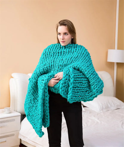 A person holds a large, chunky-knit turquoise yarn throw blanket in a bedroom.
