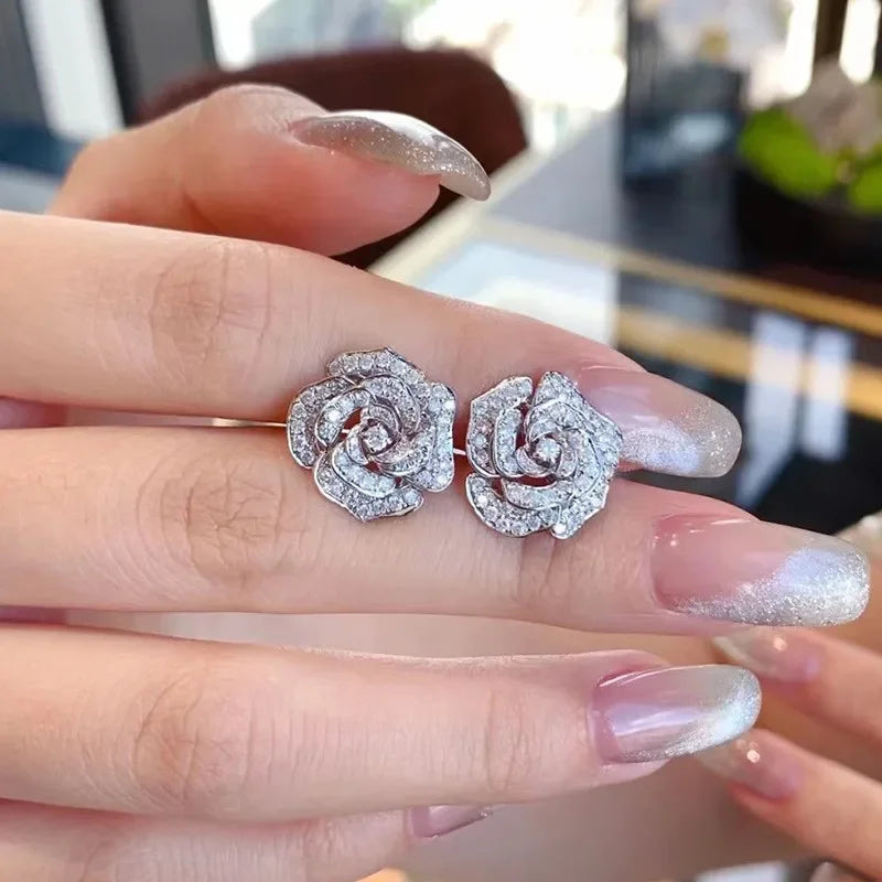 Silver-tone camellia flower stud earrings paved with sparkling clear zirconia crystals held on fingers.
